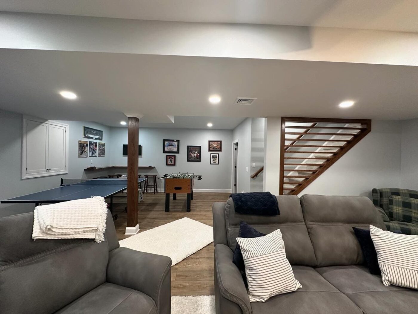 A Newly Finished Basement With Comfy Couches, Brand New Lighting, Walls, And A Stripe Design On The Staircase Wall, With An Area For Games In The Back, Such As Table Tennis And Foosball, In Lancaster County Pa